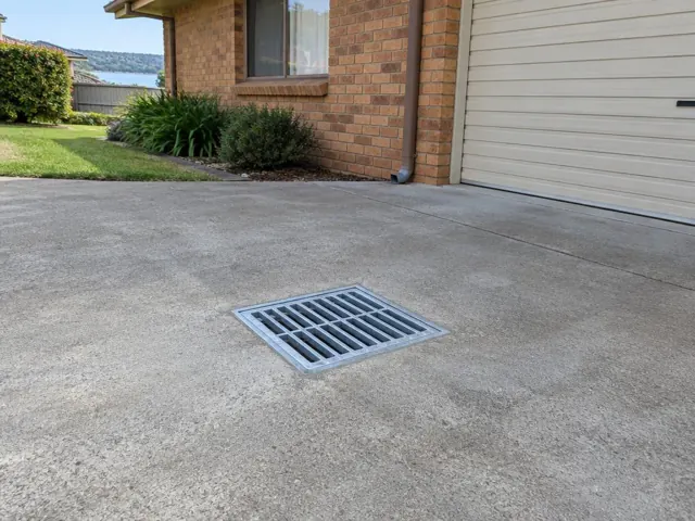 Outdoor driveway drain grate beside brick home and garage on a dry concrete driveway in Eleebana