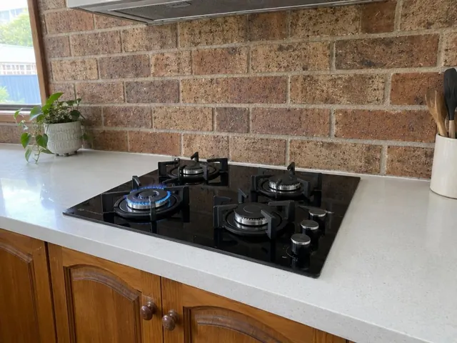 Four-burner gas cooktop set into a light benchtop with brick splashback and timber cabinets in Warners Bay